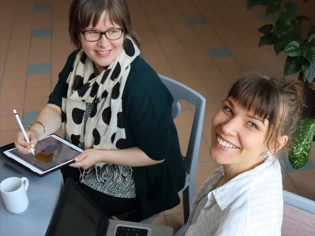 Kaksi iloista opiskelijaa opiskelemassa tietokoneella ja pädillä