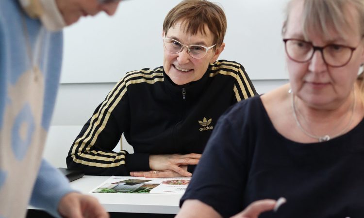 Kuvassa iloinen nainen kurkistaa kahden muun naisen takaa